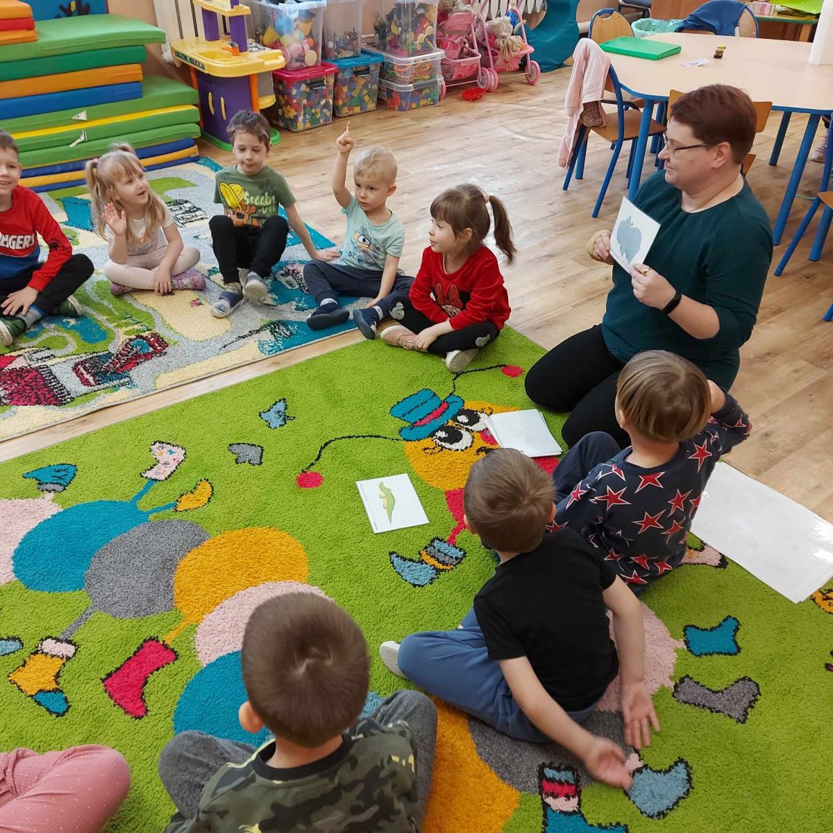 Dzieci siedzą w kółku z bibliotekarką i podnoszą ręce zgłaszając się do odpowiedzi.