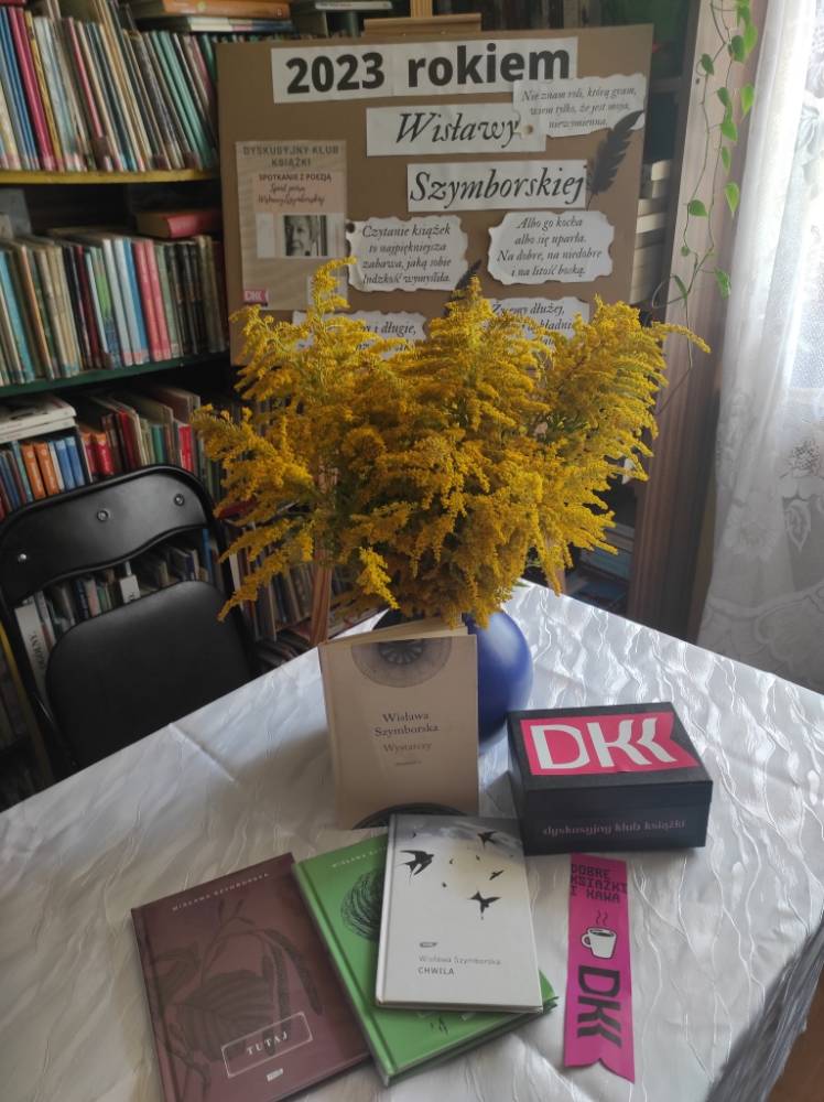 Tomiki poezji Wisławy Szymborskiej rozłożone obok dzbanka z letnimi trawami na stole w bibliotece.