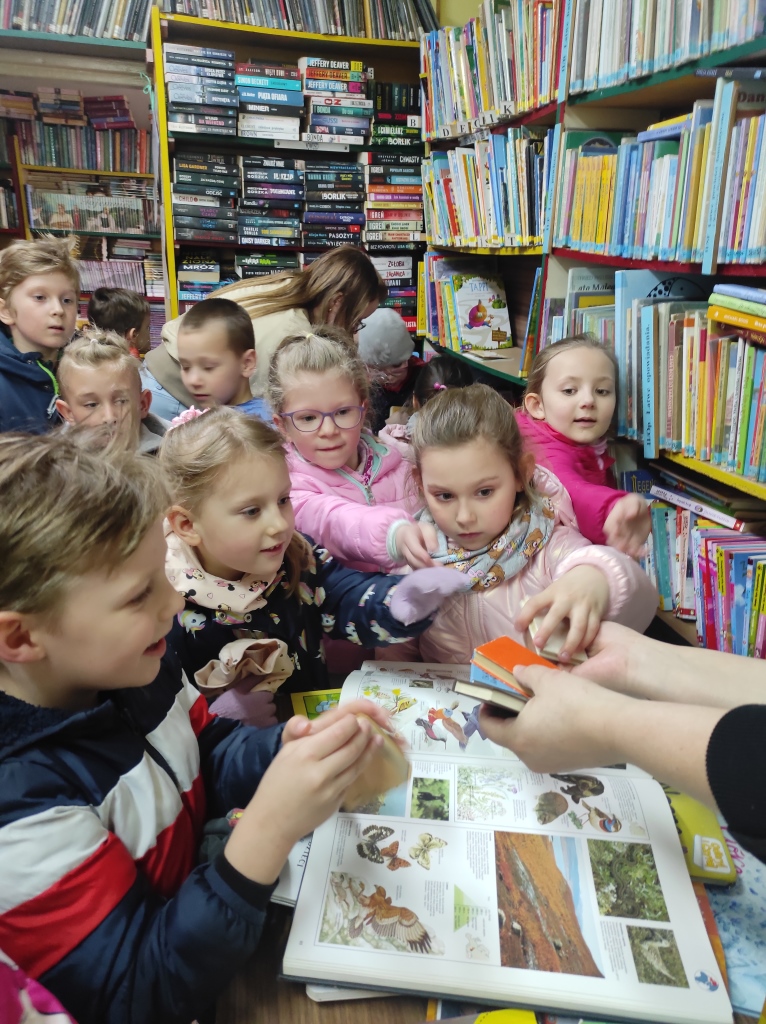 Dzieci gromadzą się przy blacie biurka bibliotekarki, żeby obejrzeć prezentowane przez nią maleńkie książeczki.